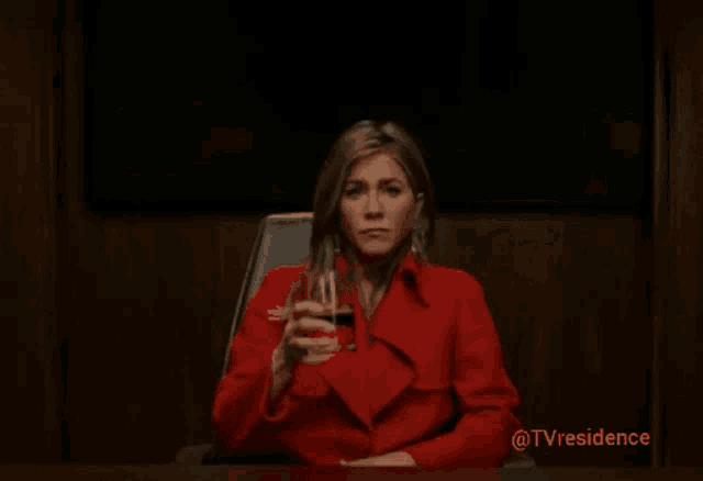 a woman in a red coat sits at a table drinking from a glass