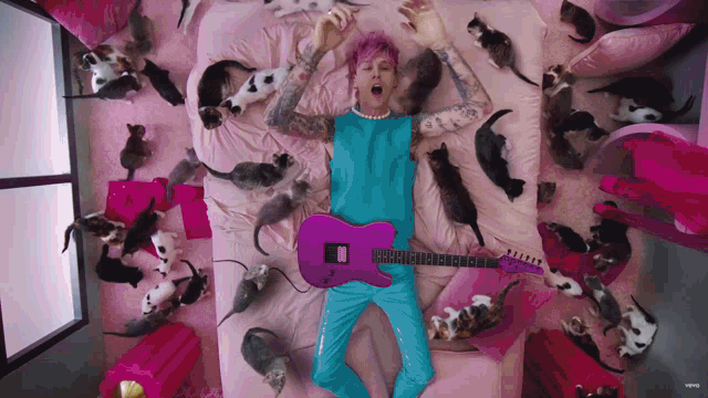 a man laying on a bed surrounded by cats holding a pink guitar
