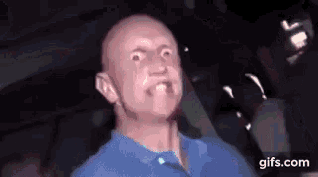 a bald man in a blue shirt is making a funny face while sitting in a car .
