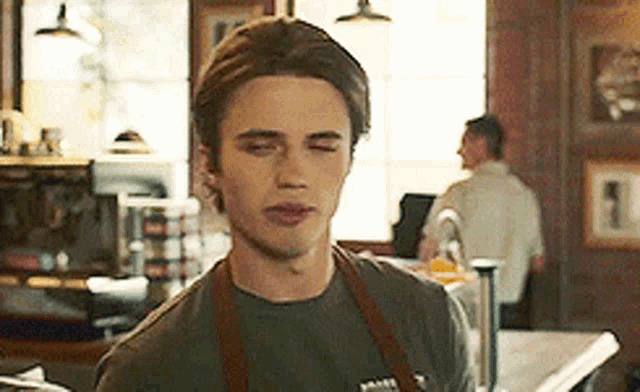 a young man wearing an apron and a t-shirt is standing in a kitchen with his eyes closed .