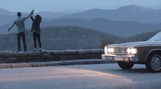 a man and a woman are standing on a wall looking at a car driving down the road