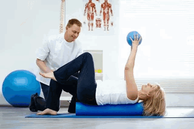 a woman is laying on a foam roller while a man holds a pilates ball .