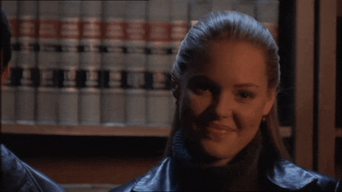 a woman in a leather jacket is smiling in front of a bookshelf with books on it