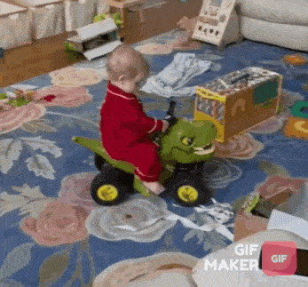 a baby in a red pajamas is riding a toy dinosaur on a floral rug .