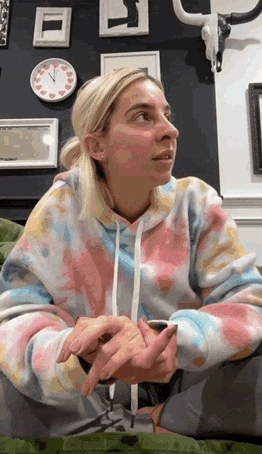 a woman wearing a tie dye sweatshirt sits on a couch with a clock on the wall behind her