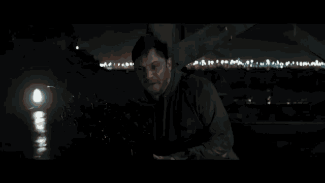 a man in a hoodie stands in the dark with a city in the background