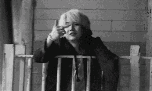 a black and white photo of a woman sitting on a porch making a peace sign with her hand .