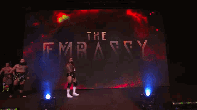 a group of wrestlers standing in front of a large screen that says the embassy