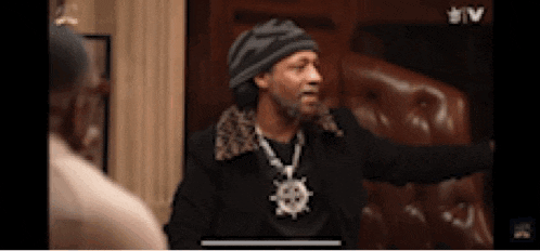 a man in a hat and necklace is sitting in a chair talking to another man .