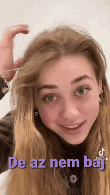 a girl with long blonde hair and blue eyes is smiling with the words de az nem baj on the bottom of her face .