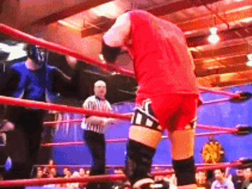 a man in a red shirt is standing in a wrestling ring with a referee behind him
