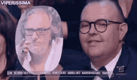 a man wearing glasses and a tuxedo is smiling in front of a picture of another man 's face .
