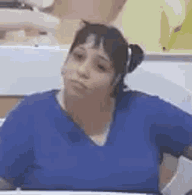 a woman in a blue scrub top is sitting at a table looking at the camera .