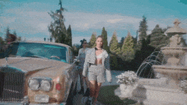 a woman in a plaid jacket and shorts is standing next to a car in front of a fountain .