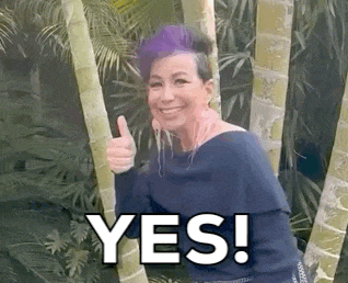 a woman with purple hair is giving a thumbs up while standing next to a bamboo tree .