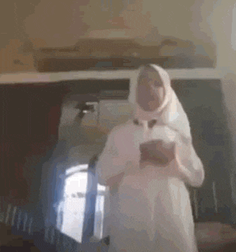 a woman in a white dress and hijab is standing in a room holding a cell phone .