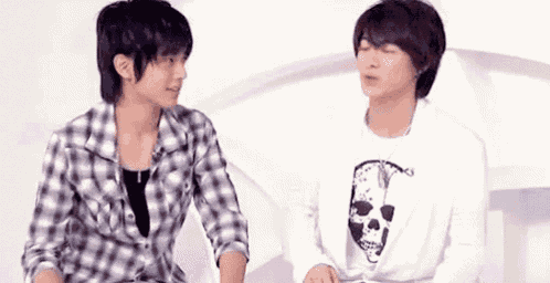 two young men are sitting next to each other and talking to each other . one of the men is wearing a skull shirt .