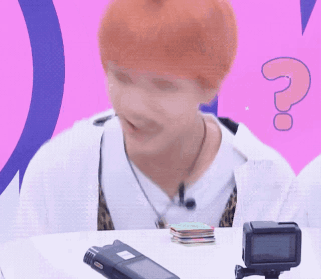 a man with red hair is sitting at a table next to a camera and a question mark ..