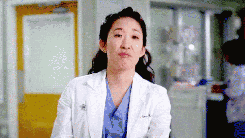 a woman in a lab coat and scrubs is making a funny face in a hospital room .
