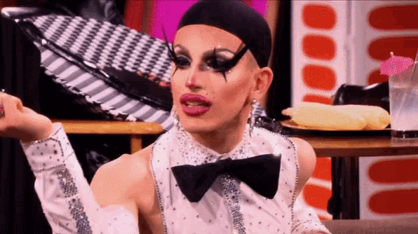 a drag queen wearing a bow tie is sitting at a table with a plate of food on it .