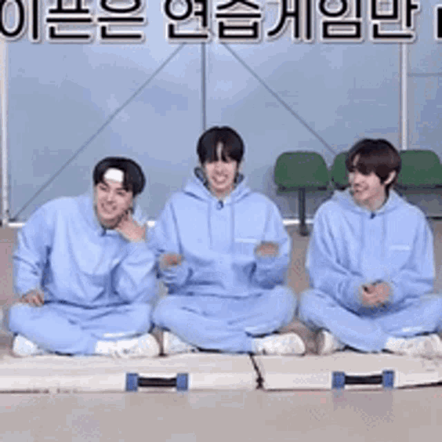three men in blue hoodies are sitting on a mat with chinese writing behind them .
