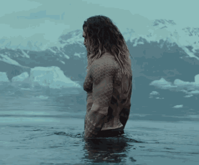 a man with long hair is standing in a body of water with mountains in the background