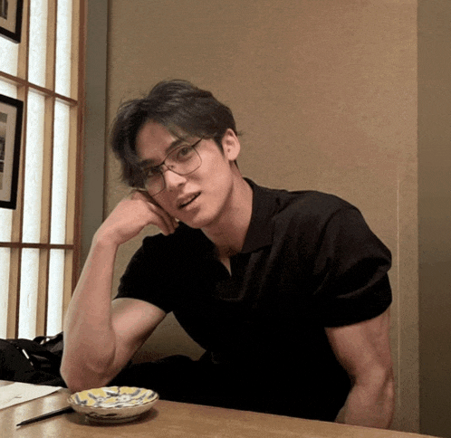 a man wearing glasses sits at a table with his hand on his face