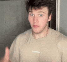 a young man in a tan t-shirt is making a funny face while standing in front of a door .