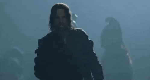 a man with long hair and a beard is standing in front of a group of people in the dark .