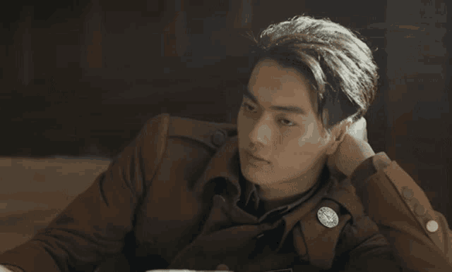 a man in a brown jacket with a badge on his chest is sitting at a table with his head resting on his hand .