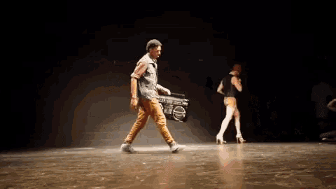 a man carrying a boombox on a stage with a woman dancing in the background
