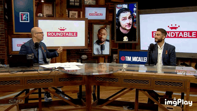 two men are sitting at a table in front of screens that say roundtable
