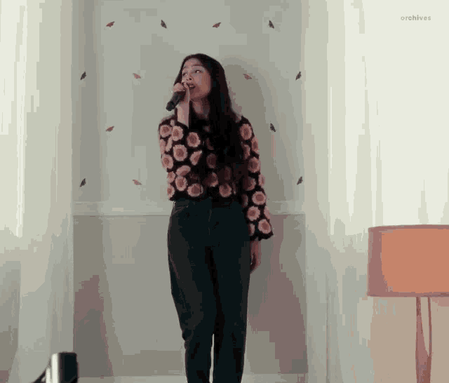 a woman is singing into a microphone in a room with a lamp .