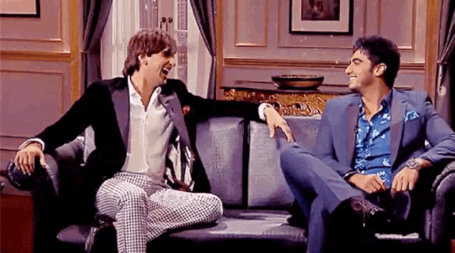two men in suits are sitting on a couch and laughing together