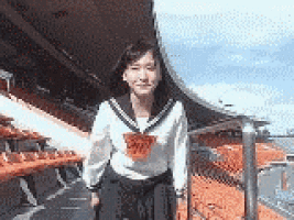 a girl in a school uniform is standing in front of a stadium with orange seats .