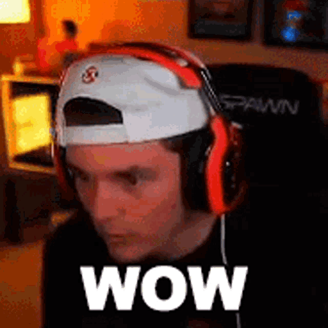 a man wearing headphones and a baseball cap is sitting in a chair with the word wow on his face .