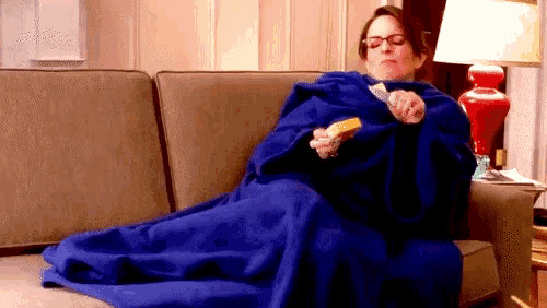 a woman wrapped in a blue blanket is sitting on a couch eating a sandwich