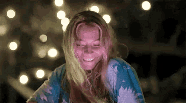 a woman with long blonde hair is smiling in front of a blurry background of lights .