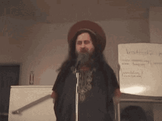 a man with long hair and a beard stands in front of a microphone in front of a white board that says " waste "