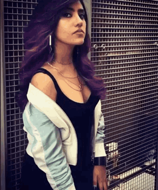 a woman with purple hair and a white jacket is standing in front of a fence .