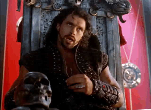 a man with long hair and a beard is sitting on a throne with a skull in front of him