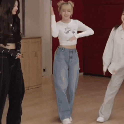 a woman in a white crop top and blue jeans is dancing in a room with other women .