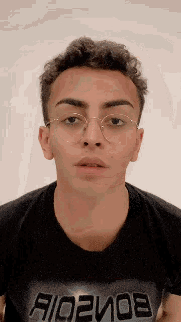 a young man wearing glasses and a t-shirt that says a/o2mo8 looks at the camera