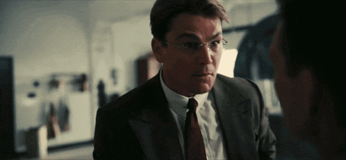 a man in a suit and tie with glasses talks to another man