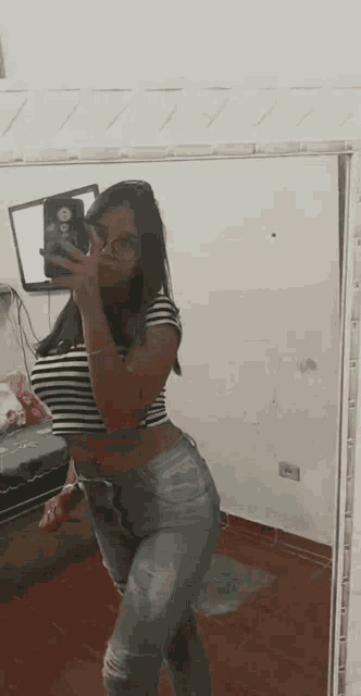 a woman is taking a selfie in front of a mirror while wearing a striped crop top and ripped jeans .