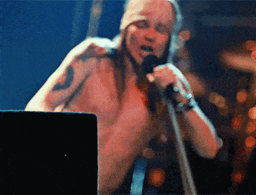 a shirtless man sings into a microphone with a tattoo on his arm