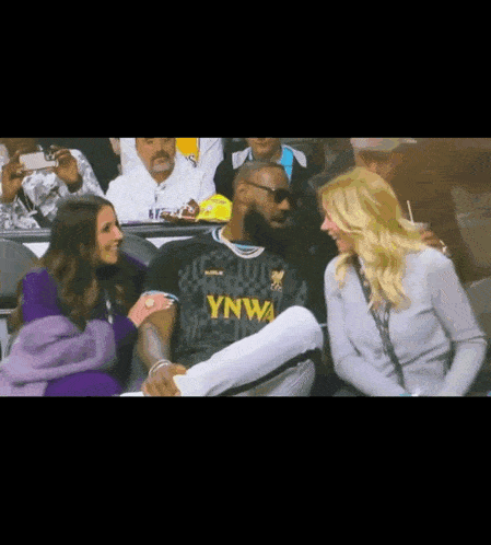 a man in a ynwa jersey sits in the stands with two women