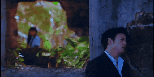 a man in a suit and tie is leaning against a wall with a woman in the background