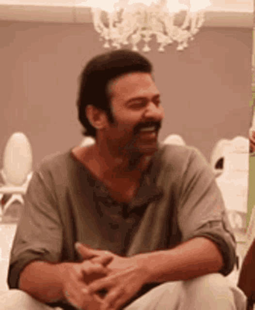 a man with a mustache is sitting on a couch with his hands folded and laughing .