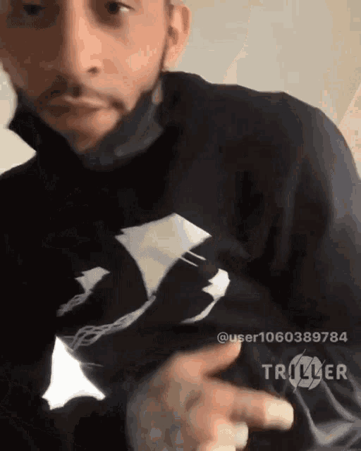 a man is wearing a black shirt with a white logo on it and a triller logo on the bottom right
